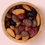 Wooden bowl filled with a mix of almonds, dried cranberries, and chocolate-dipped almonds on a pink background healthy snacking UK