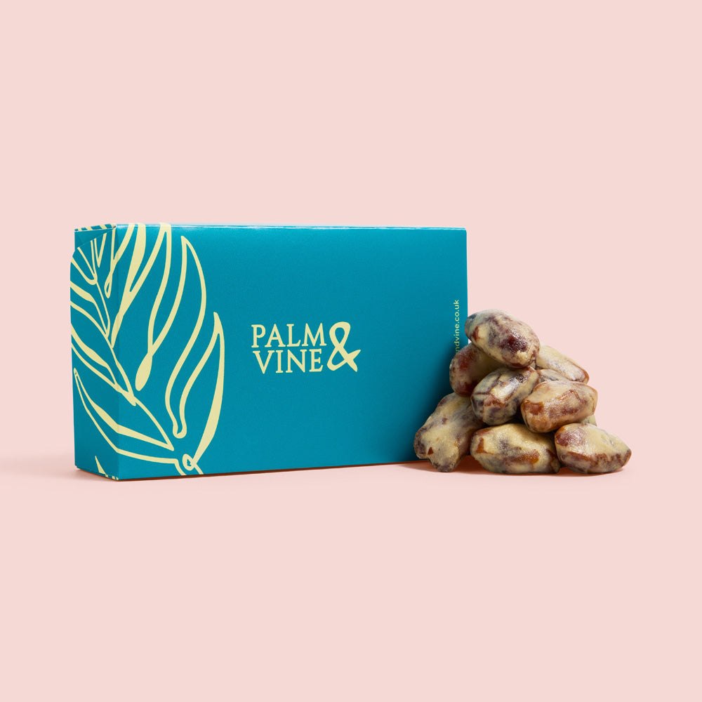 A teal and gold box of Palm & Vine peanut butter dates on a solid pale pink background.