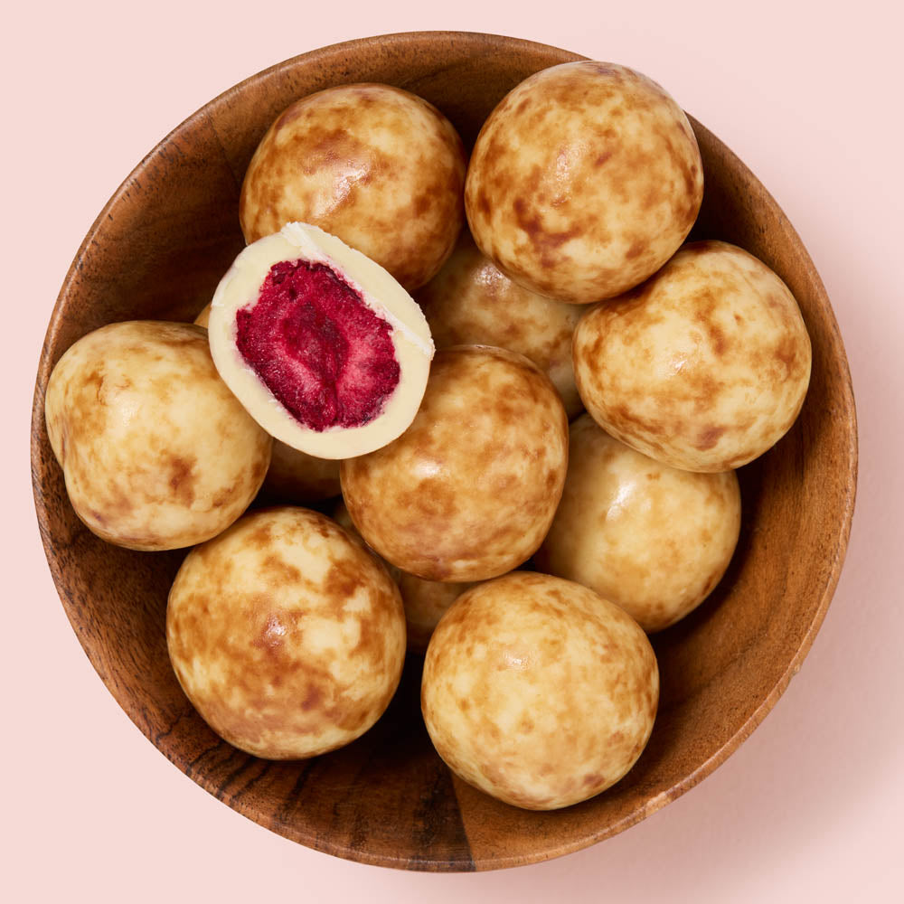 A top-down view of round, glossy beige covered cherries with brown mottling, arranged in a wooden bowl. One ball is cut open to show a bright pink-red center, set against a solid pale pink background.